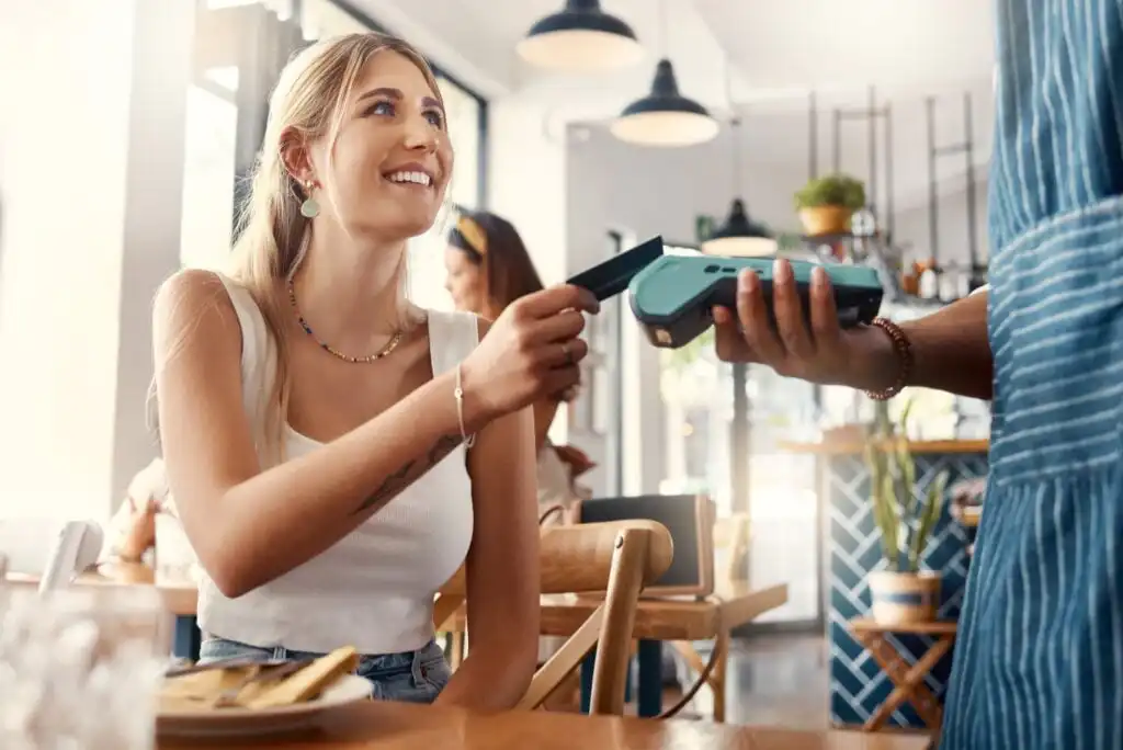 A smiling woman sitting at a cafe table pays for her meal using her smartphone, holding it towards a payment terminal presented by a person wearing a striped apron.