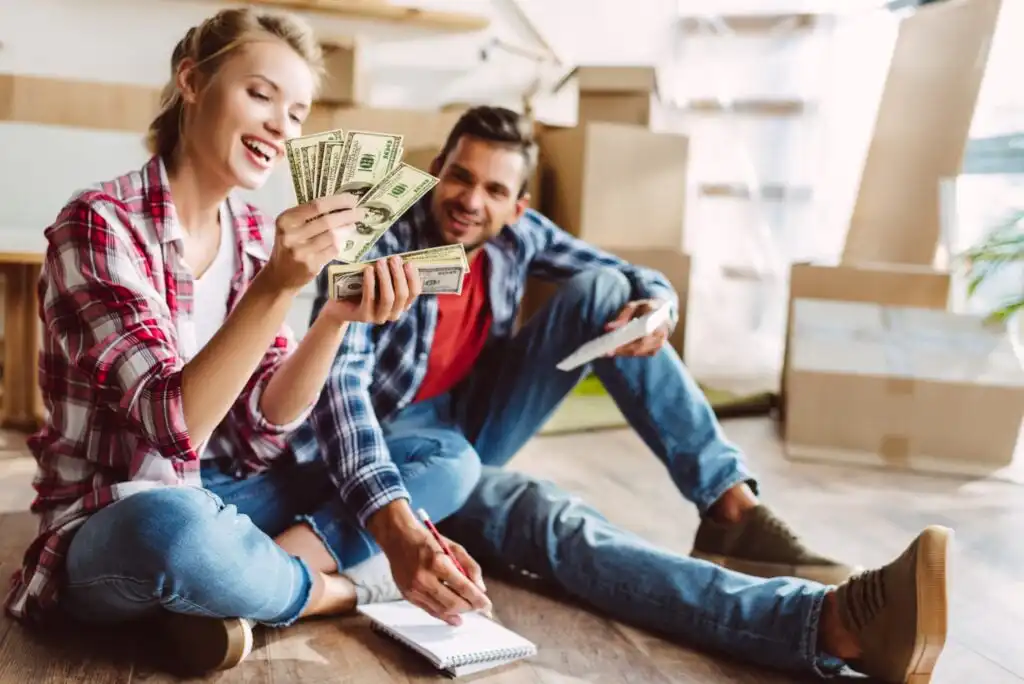 A smiling woman sits on the floor counting cash, while a man sits beside her holding paper. Both are casually dressed, surrounded by cardboard boxes, suggesting they are moving or unpacking in a new home.