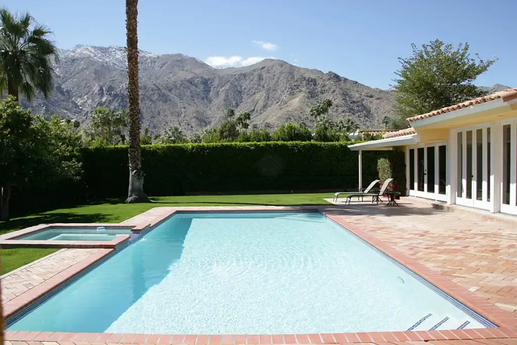 A backyard swimming pool with a hot tub, surrounded by a brick patio and lawn, with lounge chairs near a modern house; mountains and palm trees are visible in the background under a blue sky.