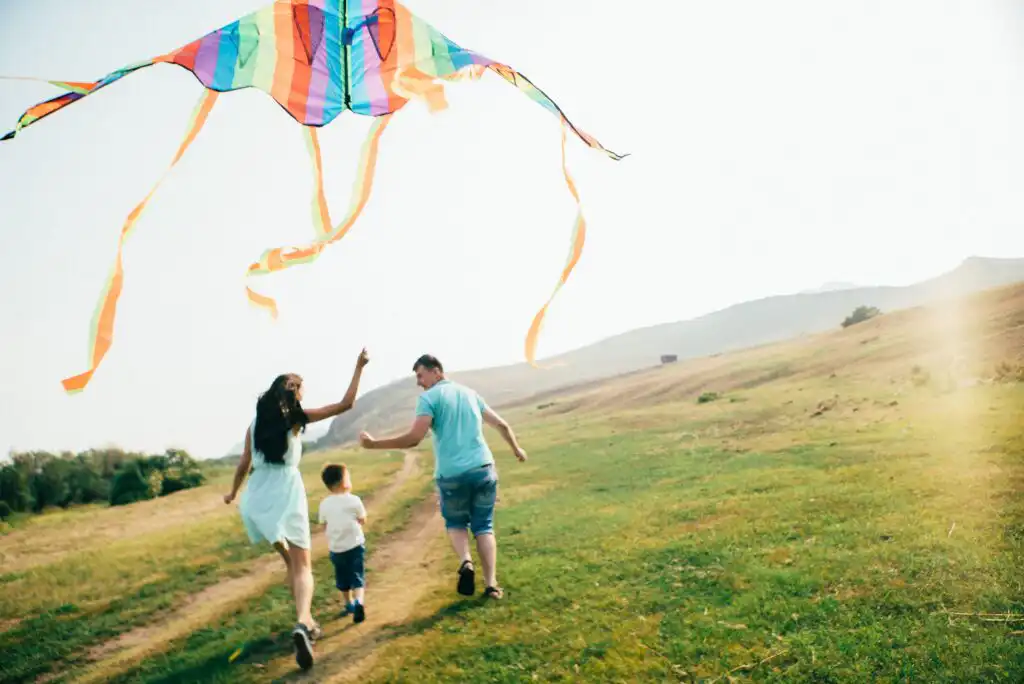 A woman, man, and young boy run together on a grassy path, flying a large, colorful kite in a sunny, open field with gentle hills in the background.