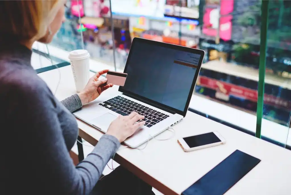 A person sitting at a desk by a window uses a laptop while holding a credit card. A coffee cup, smartphone, and tablet are on the desk, with blurred city lights visible outside.