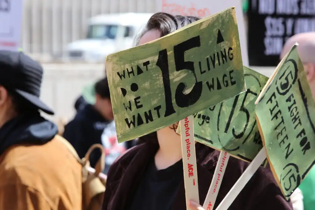 A person holds a sign reading What do we want? 15. Living wage during a protest. Other people and protest signs are visible in the background.