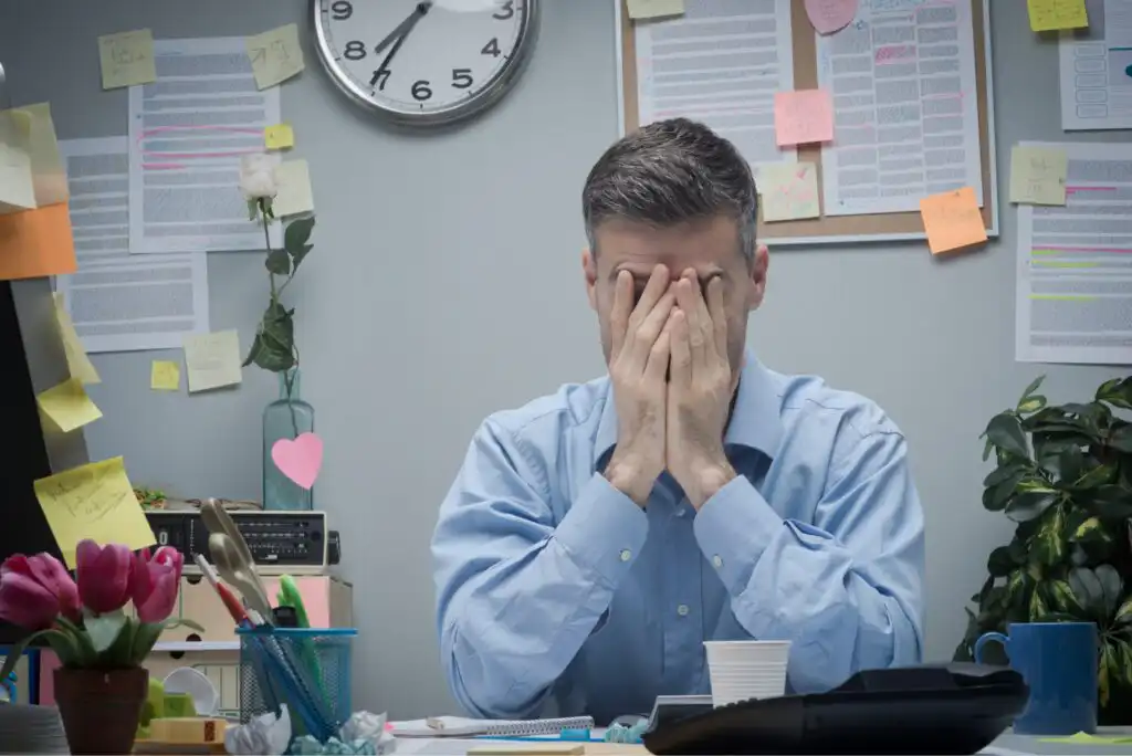 A man in a blue shirt sits at a cluttered desk with his face in his hands, appearing stressed. Papers, sticky notes, a clock, and office supplies surround him in a busy office setting.