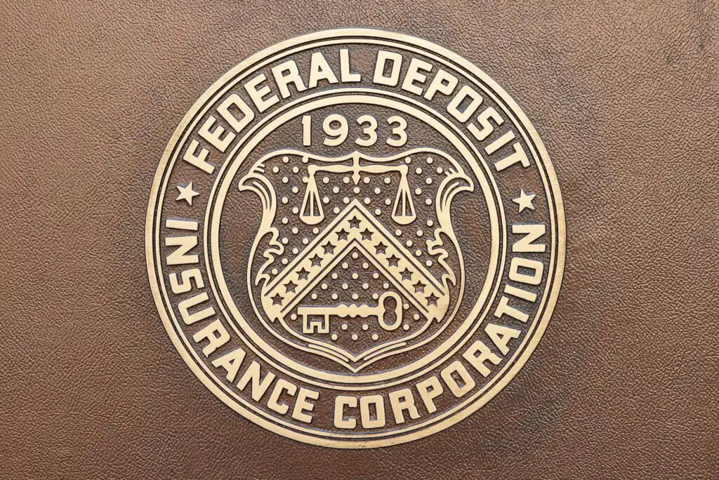 Bronze emblem of the Federal Deposit Insurance Corporation (FDIC) featuring the year 1933, a shield with a key and scales, and the organizations name in a circular design on a textured brown background.