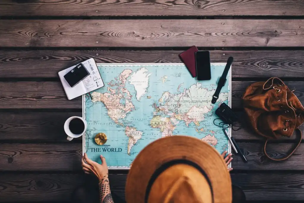 A person wearing a brown hat looks at a world map spread on a wooden surface, surrounded by a camera, phone, notebook, coffee cup, passport, and a brown bag, suggesting travel planning.