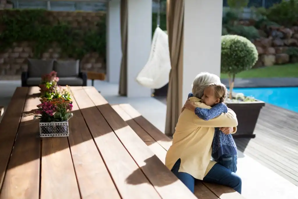 An older adult and a child sit on a wooden bench by a pool, hugging each other. The setting is an outdoor patio with flowers on the table and greenery in the background.