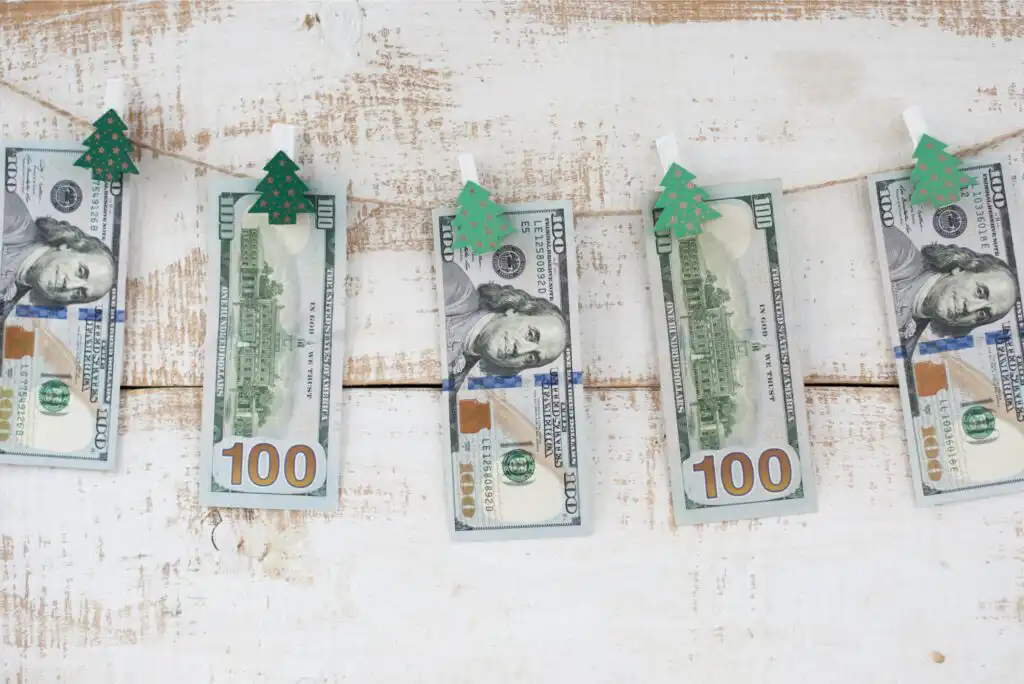 Five US $100 bills are clipped to a clothesline with small green paper Christmas tree clips, hanging against a rustic white wooden background.
