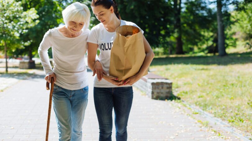 Woman Volunteer Helping Senior Citizen With Groceries Cane