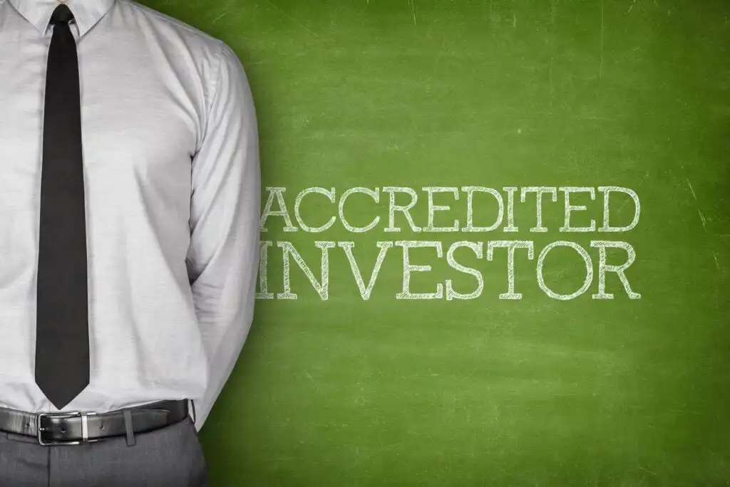 A person in a dress shirt and tie stands next to a green chalkboard with the words ACCREDITED INVESTOR written on it in white chalk.