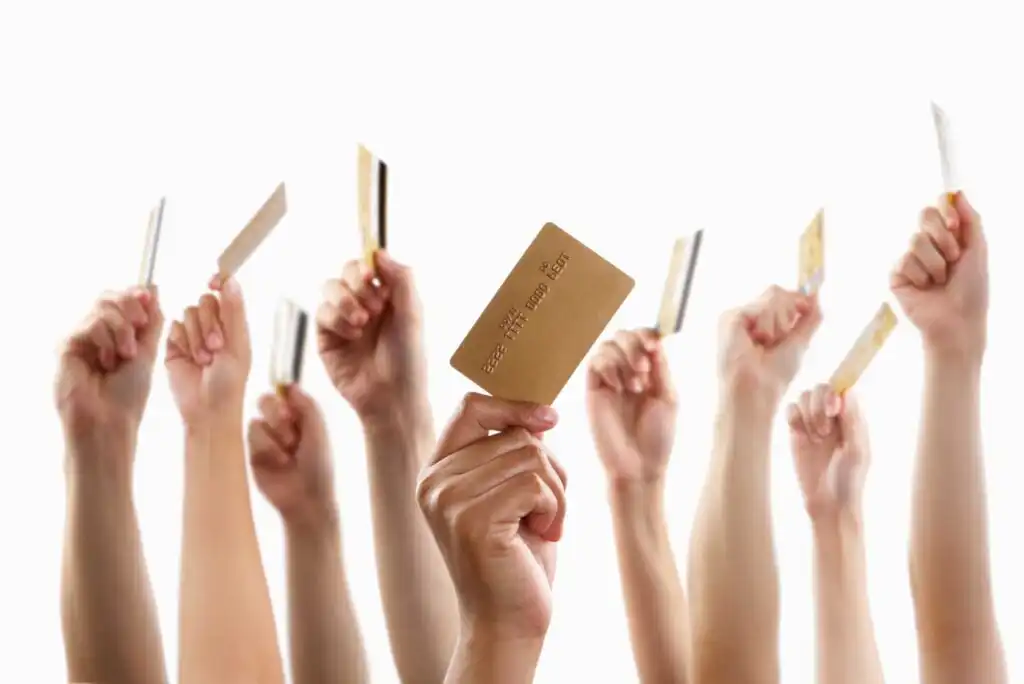 Multiple hands of diverse skin tones are raised and holding up credit or debit cards against a white background, symbolizing financial participation or spending.