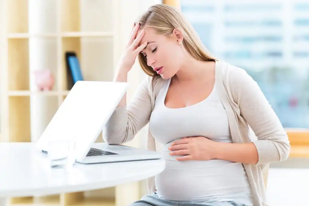 A pregnant woman sits at a table in front of a laptop, holding her belly with one hand and touching her forehead with the other, appearing stressed or tired.