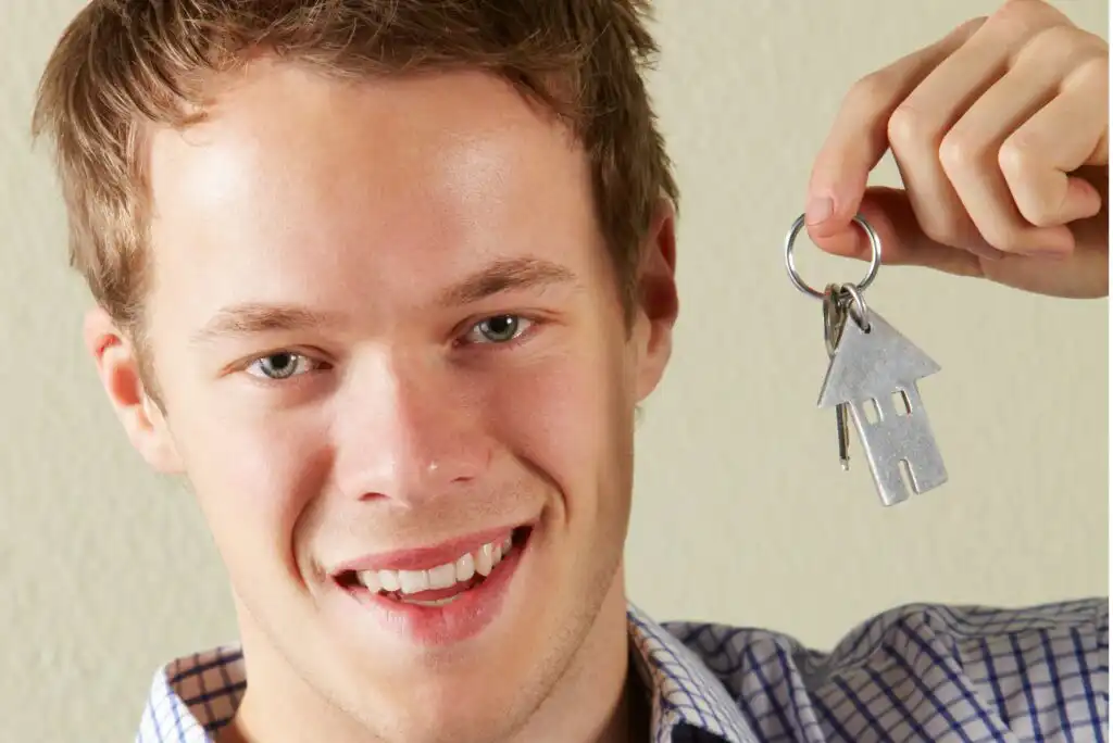A smiling young man holds up a keyring with a house-shaped keychain and keys, suggesting buying or renting a new home.