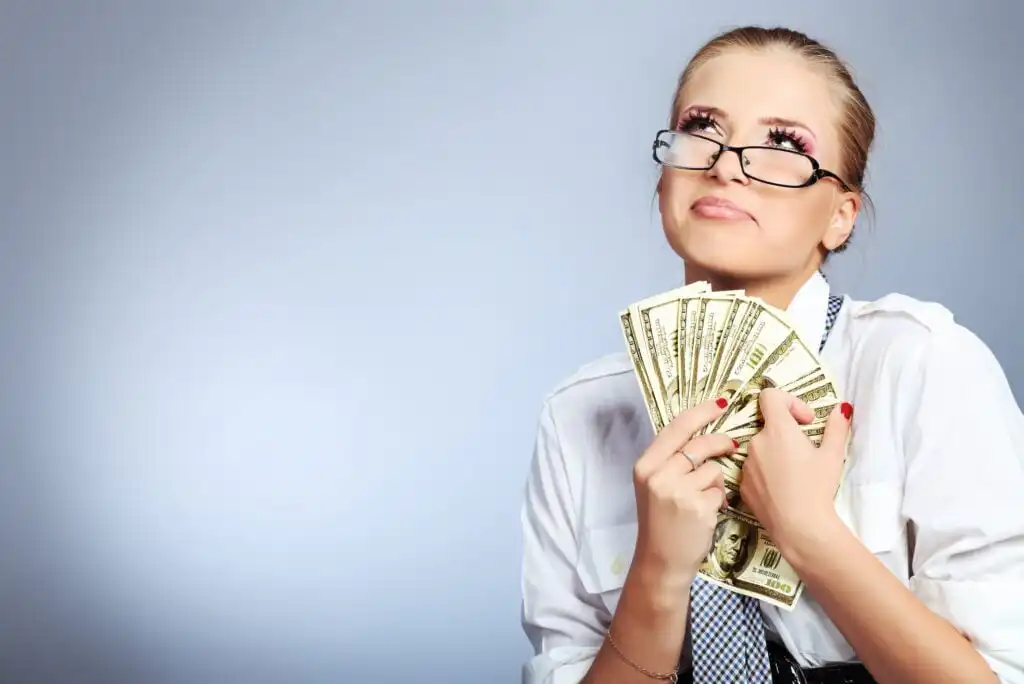 A woman wearing glasses and a white shirt holds several US dollar bills, looking upward with a thoughtful expression against a light blue background.