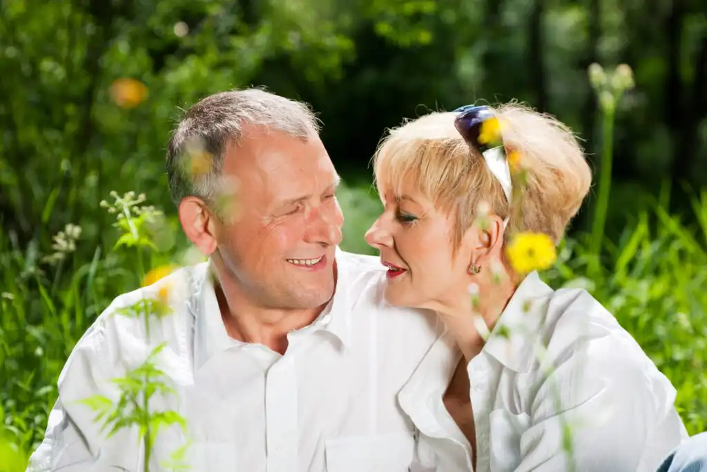 An older couple wearing white shirts sits closely together outdoors, smiling and gazing at each other, surrounded by green grass and yellow wildflowers on a sunny day.