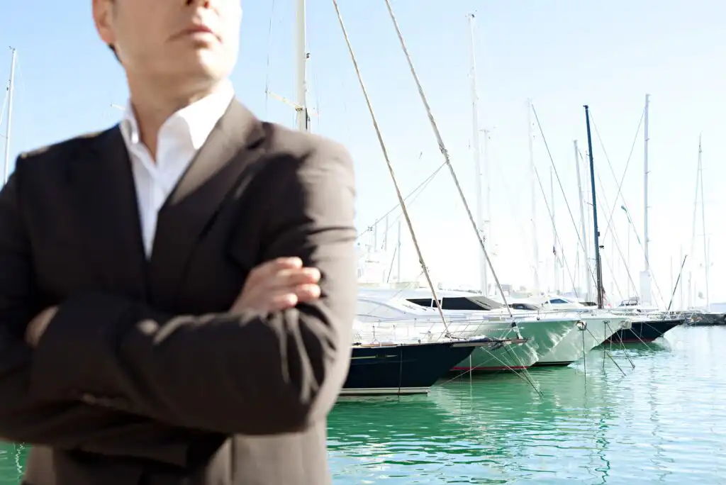 A man in a dark suit stands with arms crossed in front of a marina filled with yachts and sailboats on a sunny day. The focus is on the boats, with the man out of focus in the foreground.