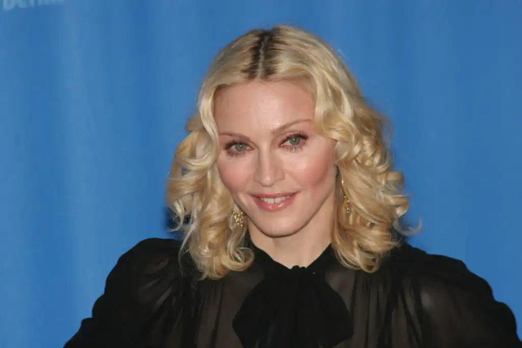A woman with shoulder-length blonde curly hair, wearing a sheer black top with a bow at the neck, smiles in front of a plain blue background.