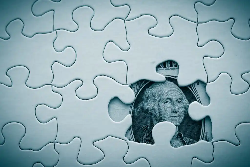 A close-up of a jigsaw puzzle with one missing piece, revealing part of a U.S. one-dollar bill showing George Washingtons face underneath the puzzle.