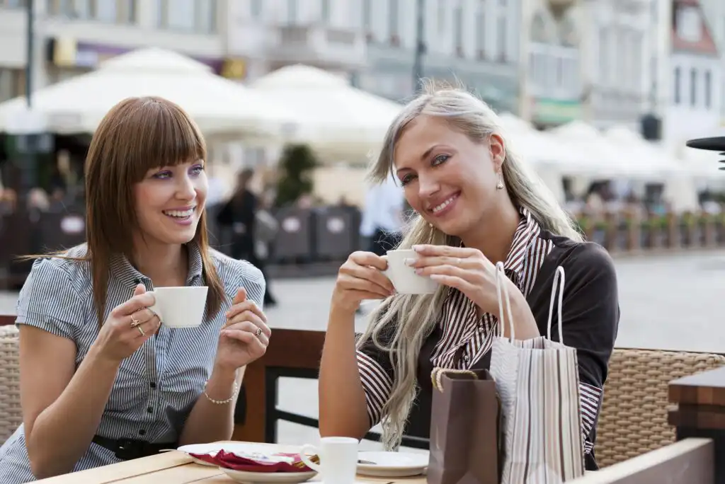Two women sitting at an outdoor café, smiling and holding coffee cups, with blurred buildings and white umbrellas in the background. Shopping bags are on the table beside them.