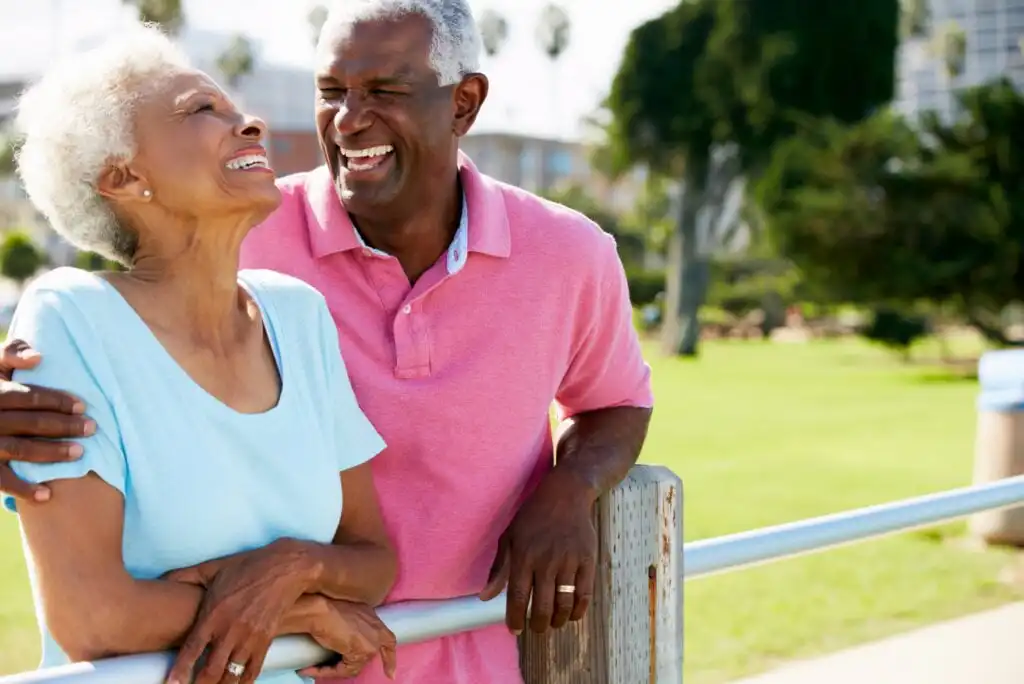 An elderly couple stands outdoors by a railing, smiling and laughing together. The woman wears a light blue shirt, and the man, in a pink shirt, has his arm around her. Green trees and sunlight are in the background.