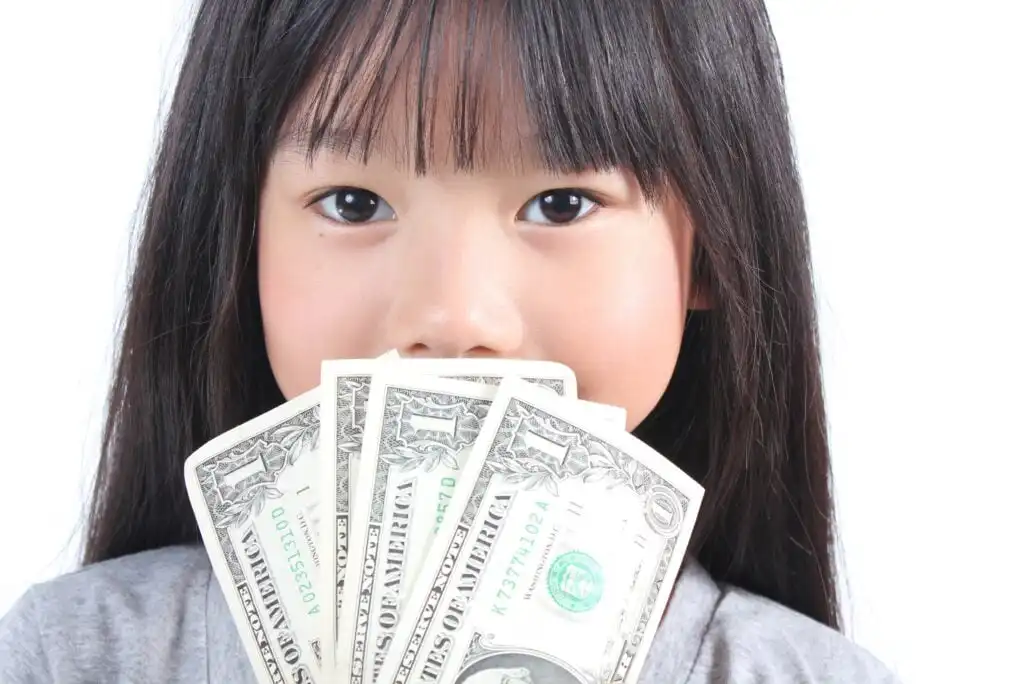 A young girl with long dark hair holds several U.S. one-dollar bills in front of her face, partially covering her mouth, with a neutral expression and a white background.
