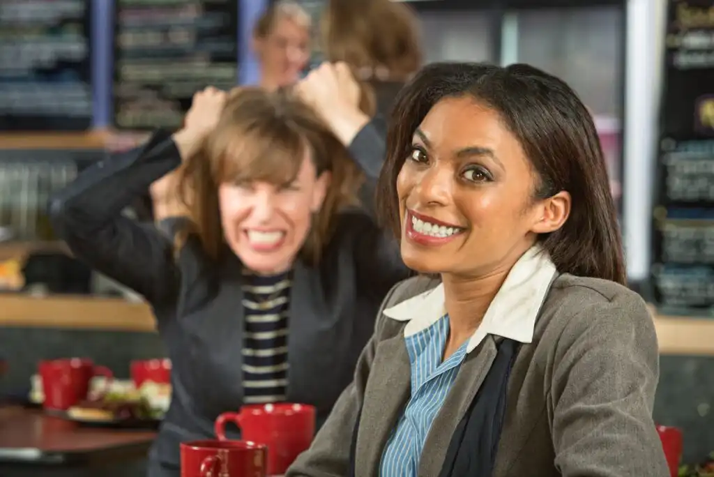 A smiling woman sits in the foreground at a cafe, while a frustrated woman in the background clenches her fists and grimaces angrily, creating a contrast in their emotions.