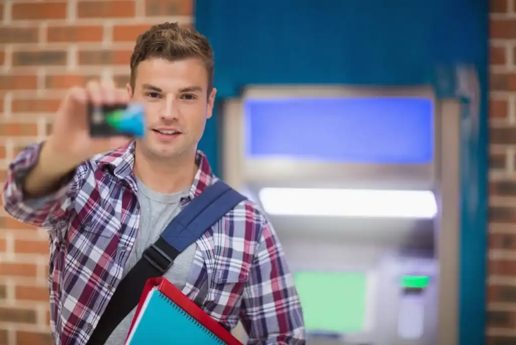 A young man holding a bank card in front of an ATM, wearing a plaid shirt and carrying notebooks and a shoulder bag, with a brick wall in the background.