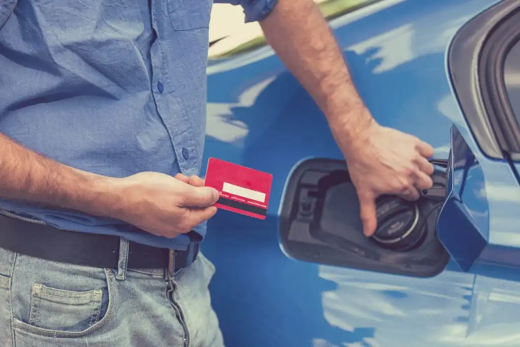 A person in jeans and a blue shirt holds a red credit card in one hand and opens the fuel tank of a blue car with the other.