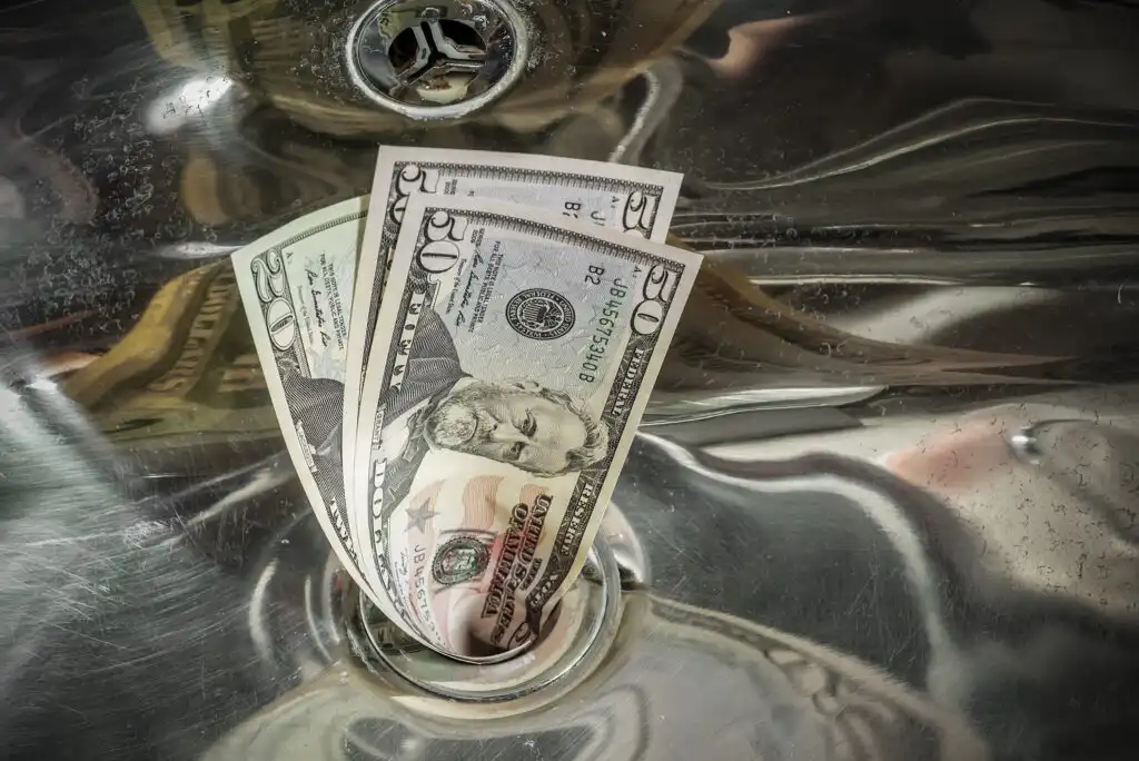 Three US dollar bills, including a $50 bill, are partially submerged in the drain of a stainless steel sink, symbolizing money being wasted or going down the drain.