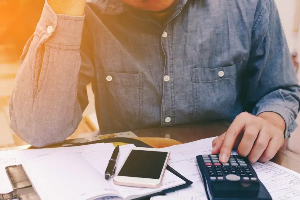A person wearing a denim shirt uses a calculator at a desk with papers, a planner, a pen, and a smartphone, appearing to concentrate or feel stressed.