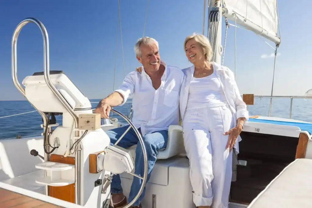 An older couple dressed in white smiles and enjoys a sunny day while sitting together on a sailboat, with blue sky and calm sea in the background.