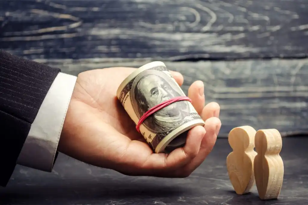 A hand in a suit holds a rolled bundle of U.S. $100 bills secured with a rubber band. Beside the hand are two small wooden human-shaped figures on a dark surface.
