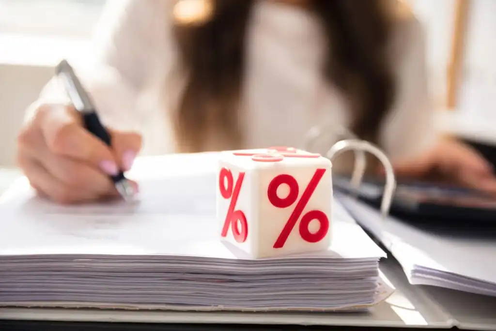 A cube with red percentage signs sits on top of a stack of documents, while a person in the background writes with a pen.