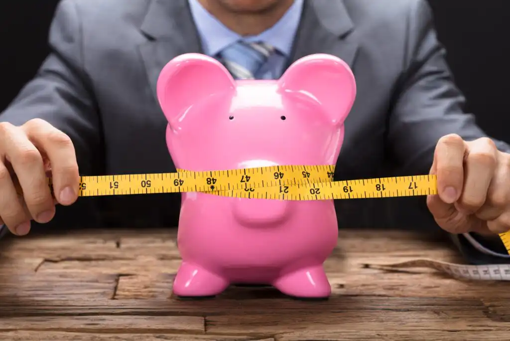 A person in a suit measures a pink piggy bank with a yellow measuring tape on a wooden table, symbolizing financial management or budgeting.