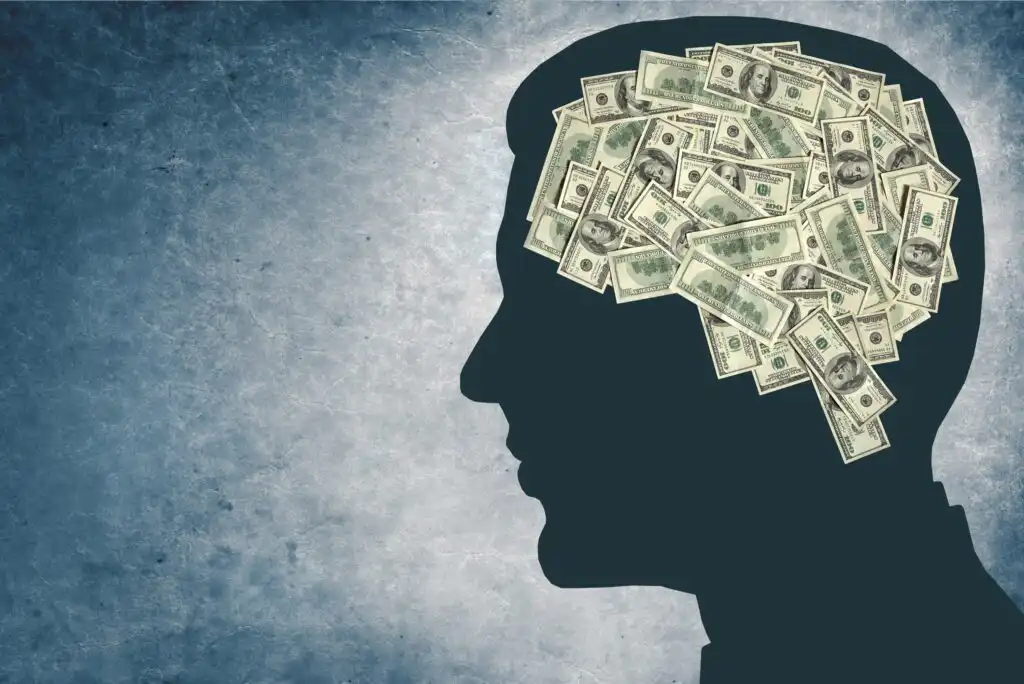 Silhouette of a person’s head filled with stacks of U.S. dollar bills, symbolizing thoughts or ideas about money. The background is textured in shades of blue and gray.