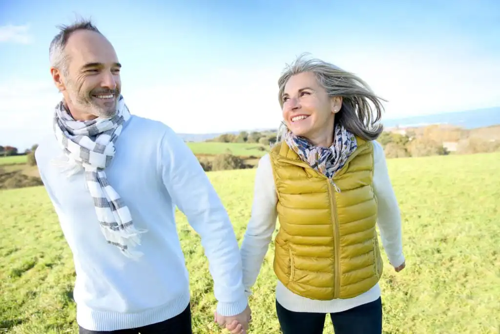 A smiling older couple, wearing warm clothes and scarves, walk hand in hand through a grassy field on a bright, sunny day.
