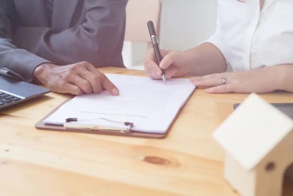 Two people sit at a wooden table; one points to a document on a clipboard while the other signs it. A laptop and a small house model are also on the table.