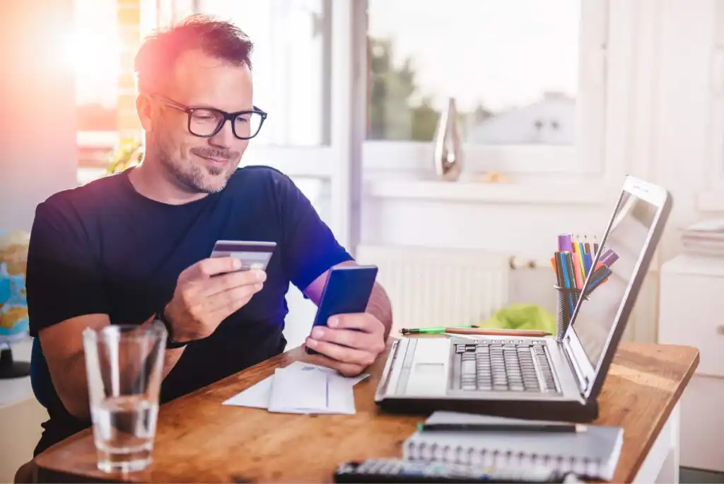 Smiling man wearing glasses sits at a table with a laptop, holding a credit card and looking at his smartphone. There are papers, a glass of water, and office supplies on the desk. Sunlight shines through the window.