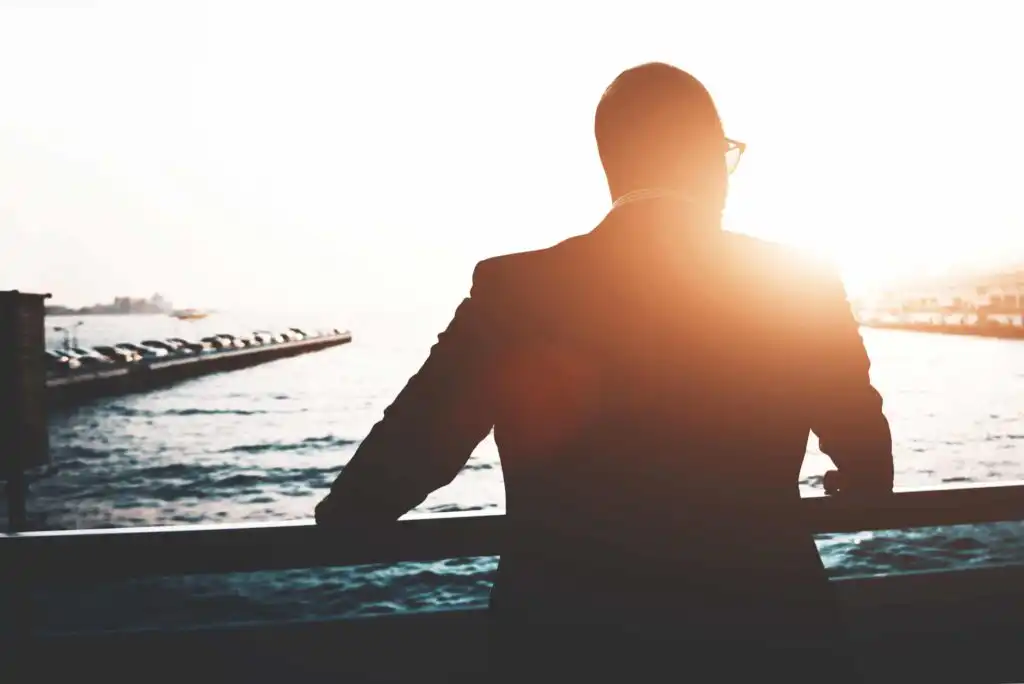 A man in a suit stands by a railing, looking out over the water at sunset. The sunlight creates a warm glow and silhouettes his figure, with boats and a pier visible in the background.