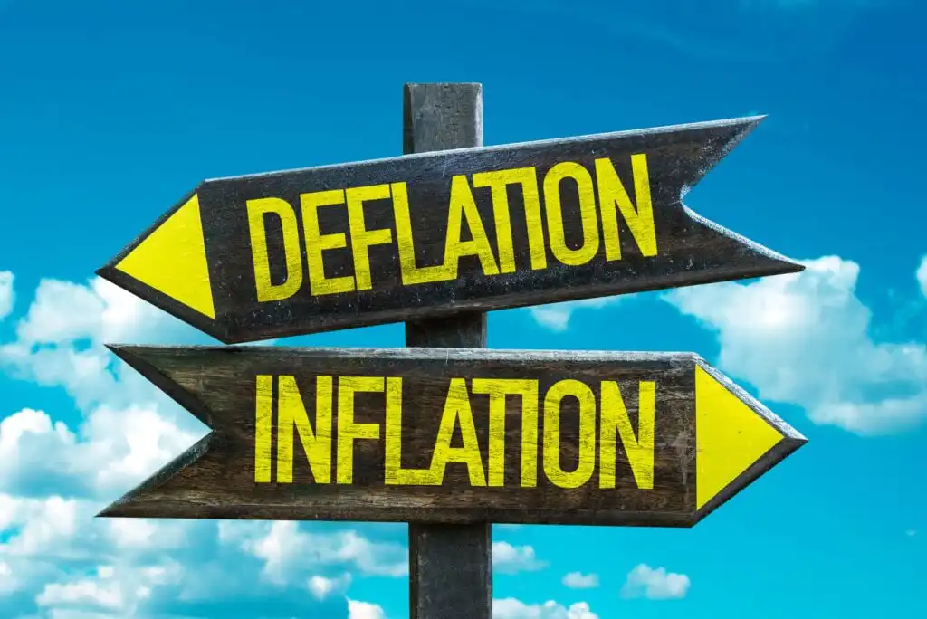 Two wooden arrow signs on a post point in opposite directions, one labeled “DEFLATION” and the other “INFLATION,” against a blue sky with scattered clouds.