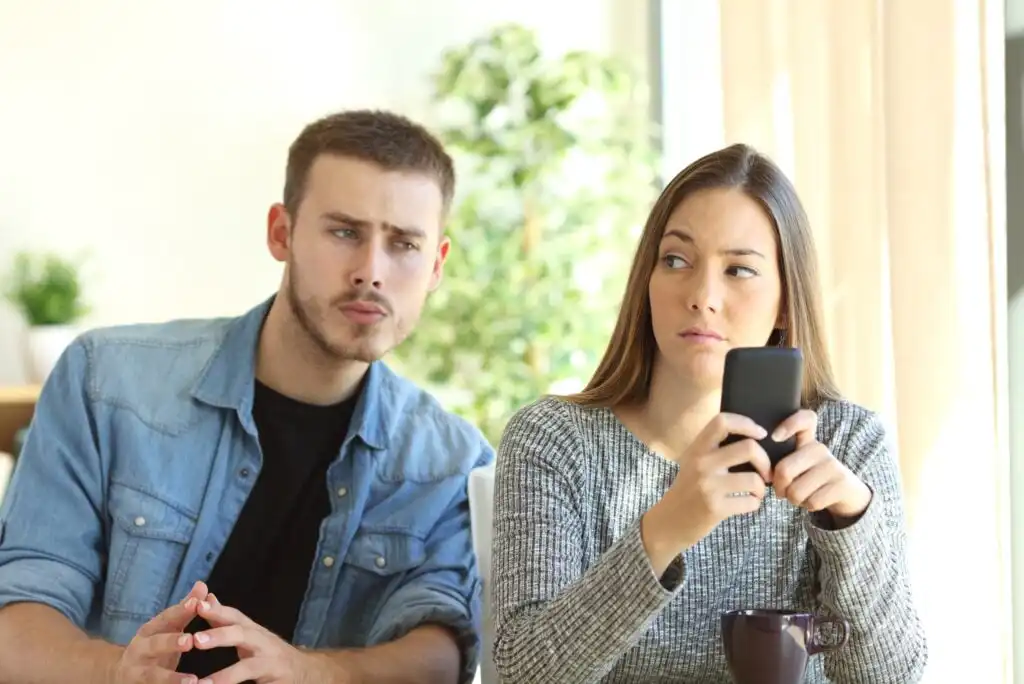 A man looks suspiciously at a woman holding a smartphone, while the woman glances back at him with a guarded expression. They are sitting indoors, and the woman also has a coffee mug.