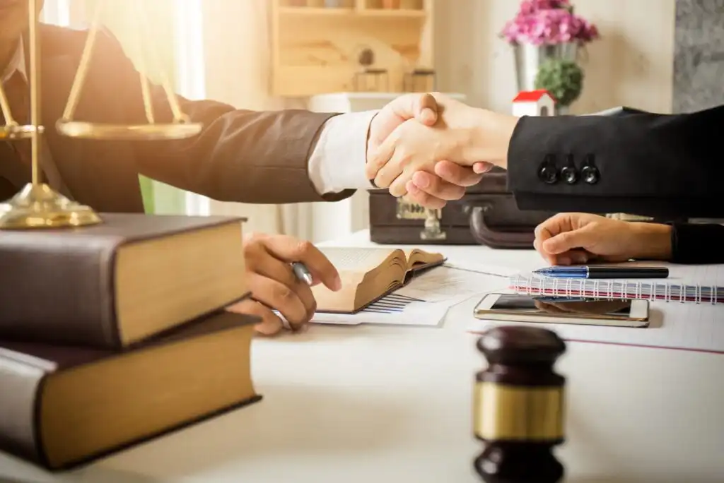 Two people in business attire shake hands across a desk with legal books, a gavel, scales of justice, a notepad, and a smartphone, suggesting a legal agreement or consultation in an office setting.