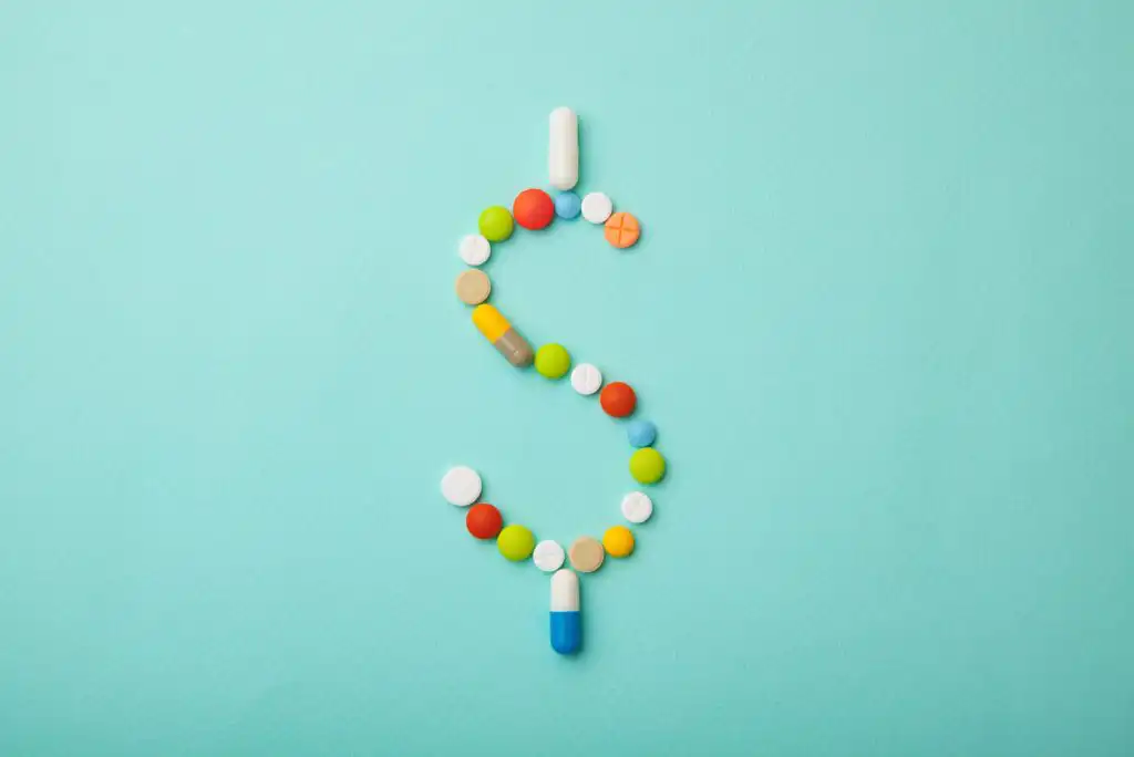 Various colorful pills and tablets arranged in the shape of a dollar sign on a light blue background.