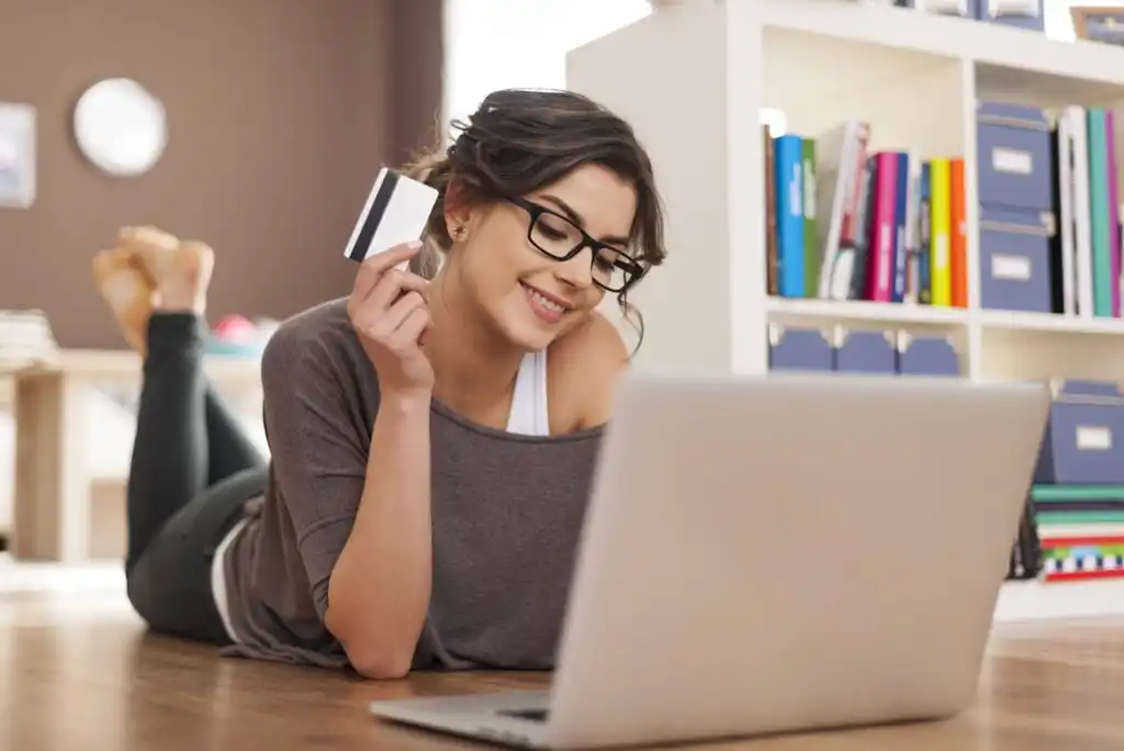 A woman with glasses lies on the floor, smiling at a laptop while holding a credit card, suggesting online shopping or payment. Bookshelves and folders are visible in the background.