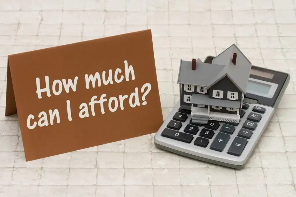 A small model house sits on a calculator next to a brown card that reads How much can I afford? on a tiled surface, suggesting a question about home affordability or budgeting.
