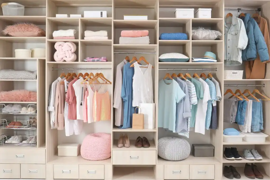 A neatly organized closet with shelves, drawers, and hanging rods displaying pastel-colored clothes, hats, bags, shoes, and folded linens. Decorative pillows and storage boxes are arranged on the shelves and floor.