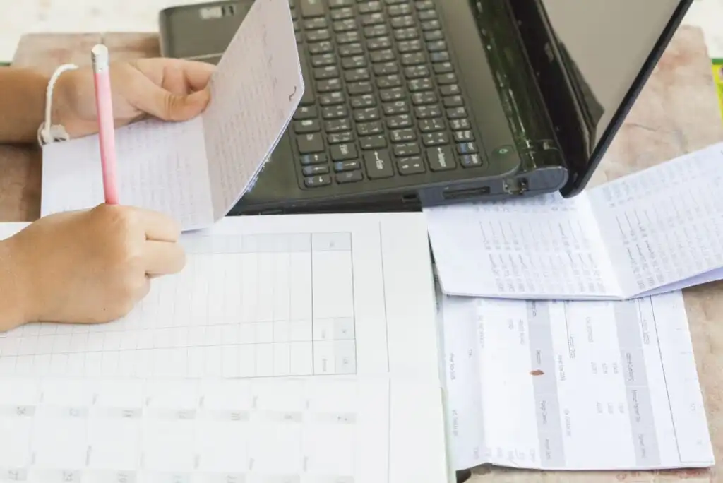 A person is holding papers while working at a desk with a laptop, documents, and a calendar spread out, appearing to organize or review information.
