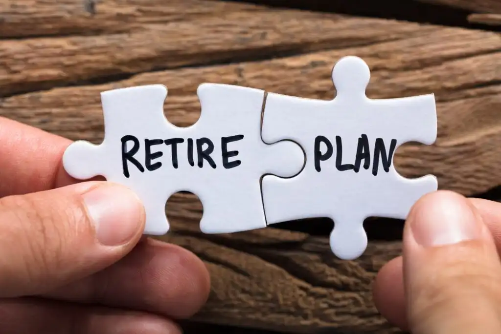 Two hands hold matching puzzle pieces together; one piece says RETIRE and the other says PLAN, symbolizing the concept of retirement planning. The background is wooden.