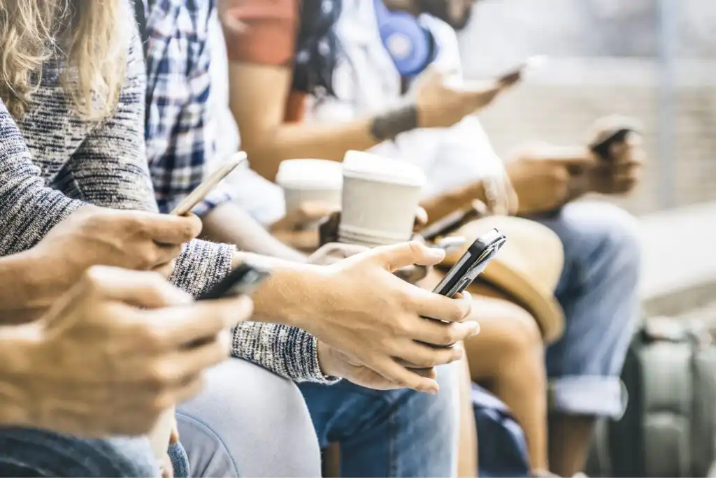 A group of people sitting side by side, holding smartphones and coffee cups, with their faces out of focus; most are looking down at their phones.