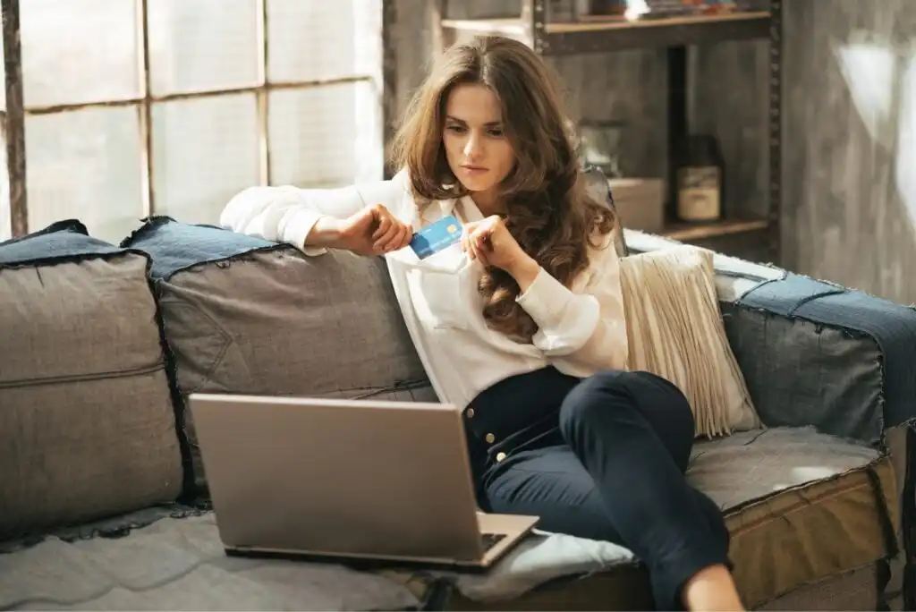 A woman sits on a sofa holding a credit card and using a laptop, appearing focused. She is dressed in a white blouse and dark pants, with sunlight streaming through large windows behind her.