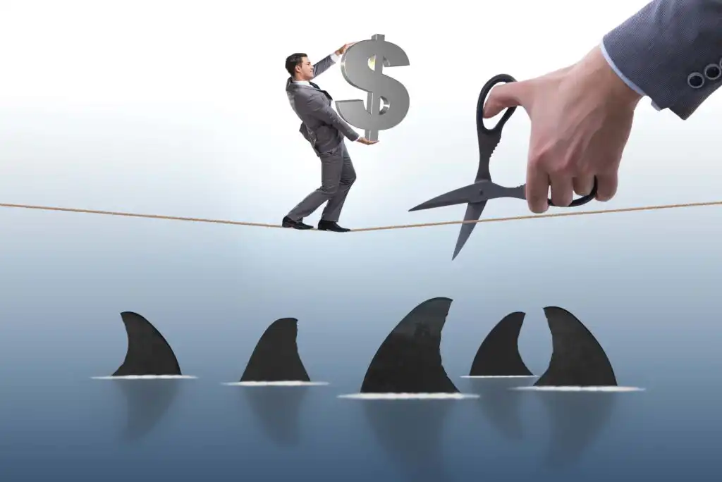 A man in a suit holding a large dollar sign walks on a tightrope over shark fins in the water, while a giant hand with scissors prepares to cut the rope.